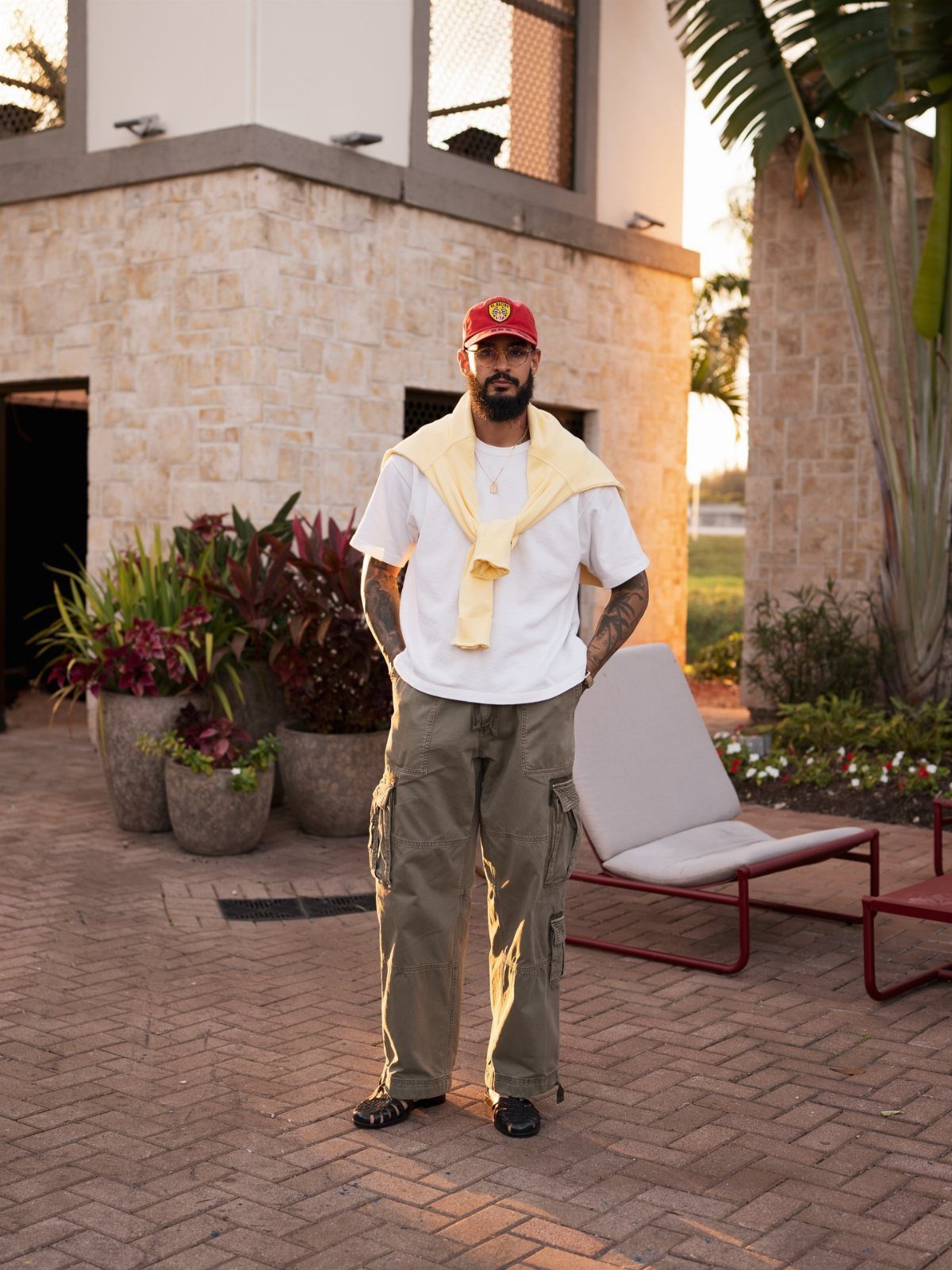 Man in white t-shirt, olive cargo pants, and red cap standing on a brick patio.