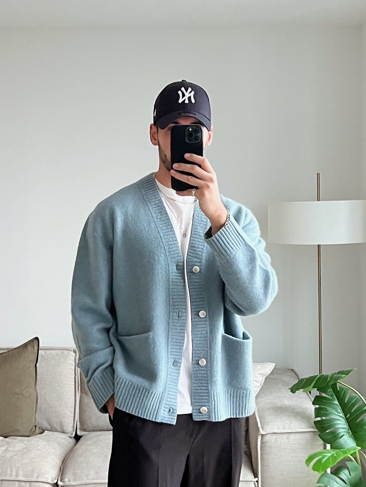 Man in a light blue chunky knit cardigan and navy baseball cap taking a mirror selfie.