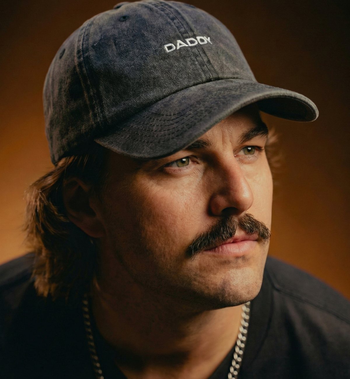 Man with a mustache wearing a dark grey baseball cap with DADDY embroidered on it.