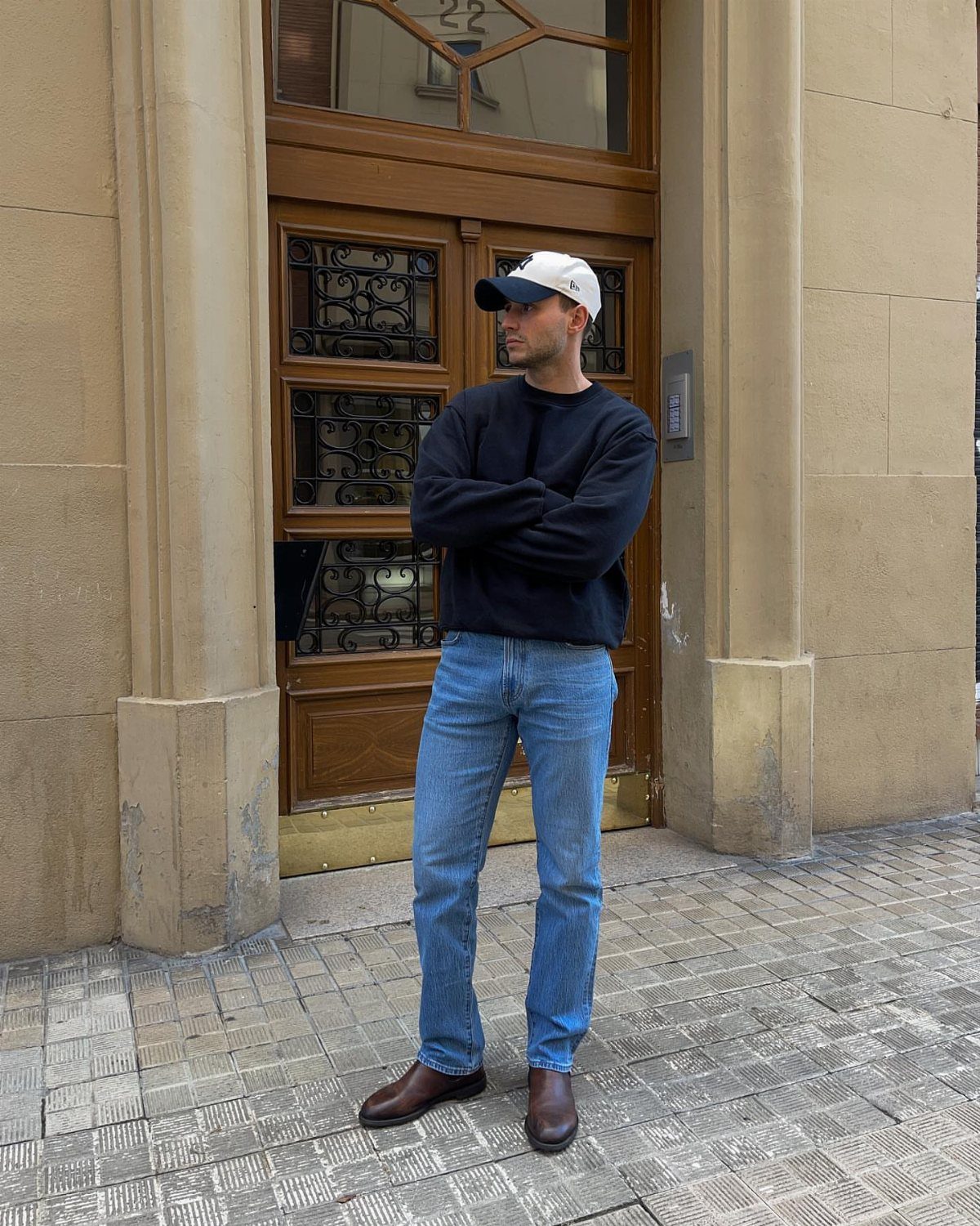 Man wearing a black sweatshirt, light blue jeans, and a baseball cap standing outdoors.