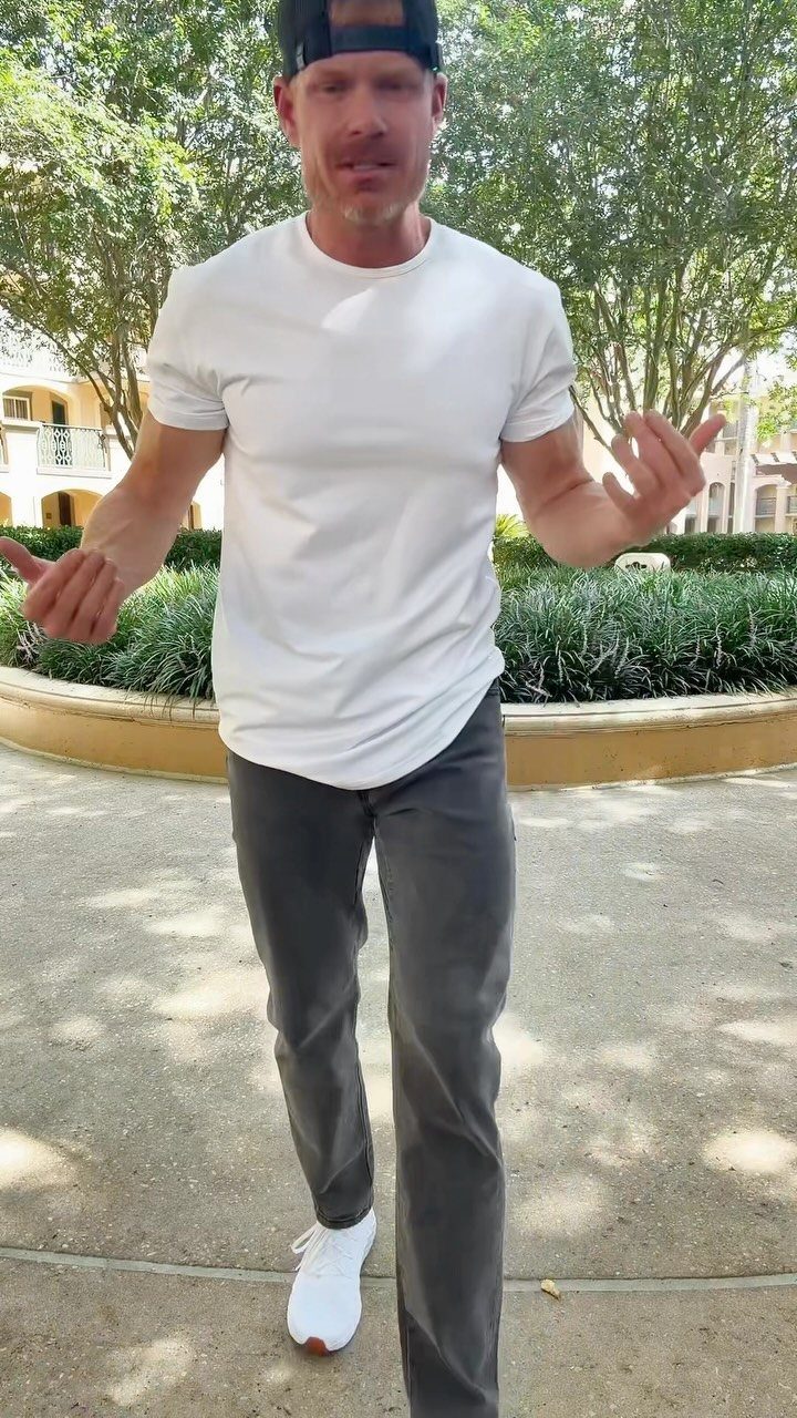 Man in a white t-shirt and dark grey trousers standing outdoors near green trees and a planter.