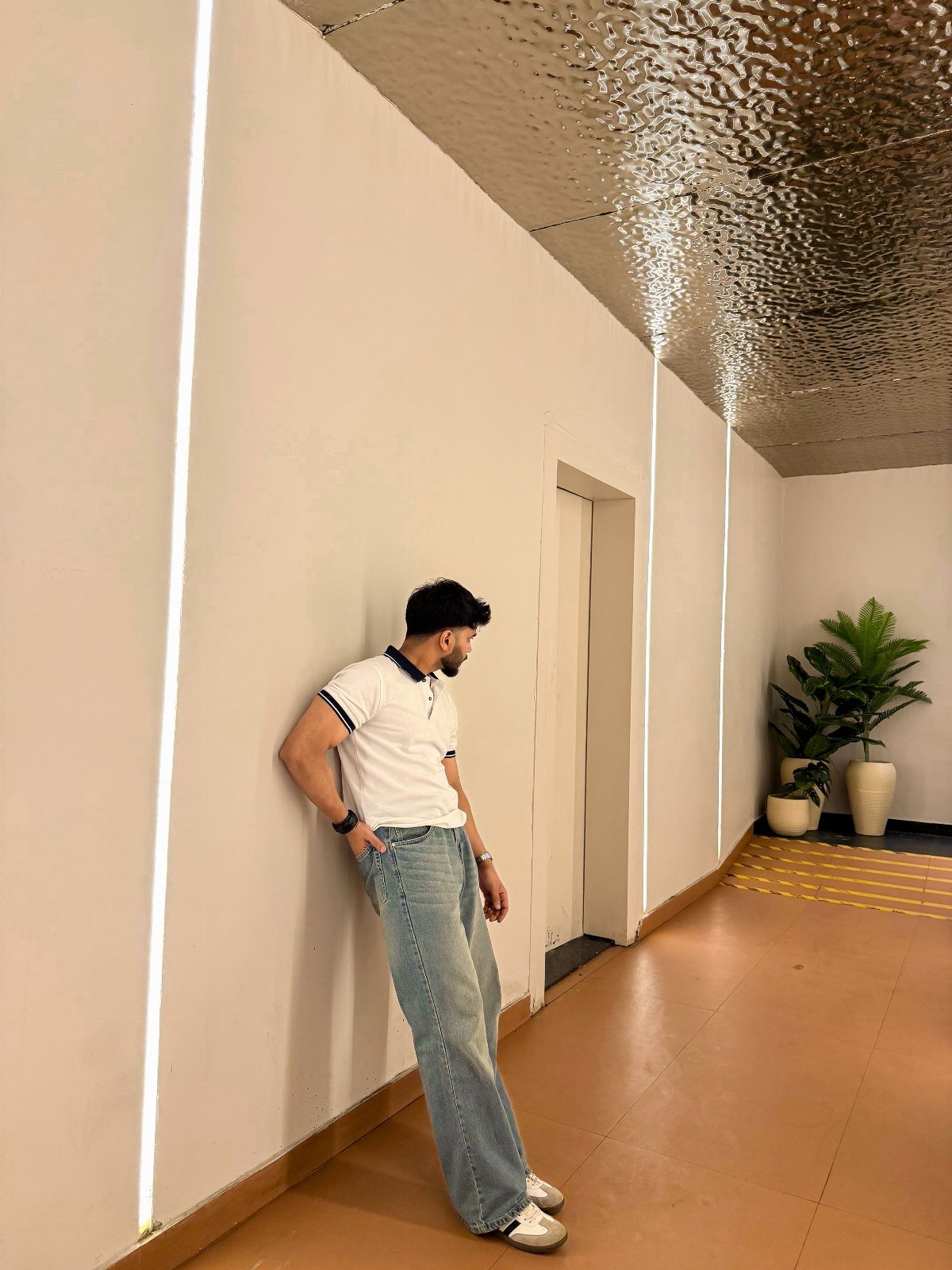 A man in a white polo and light blue jeans leaning against a white wall.