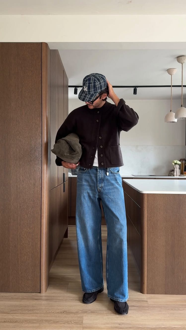 A person wearing wide-leg denim jeans, a dark jacket, and a plaid cap in a kitchen.