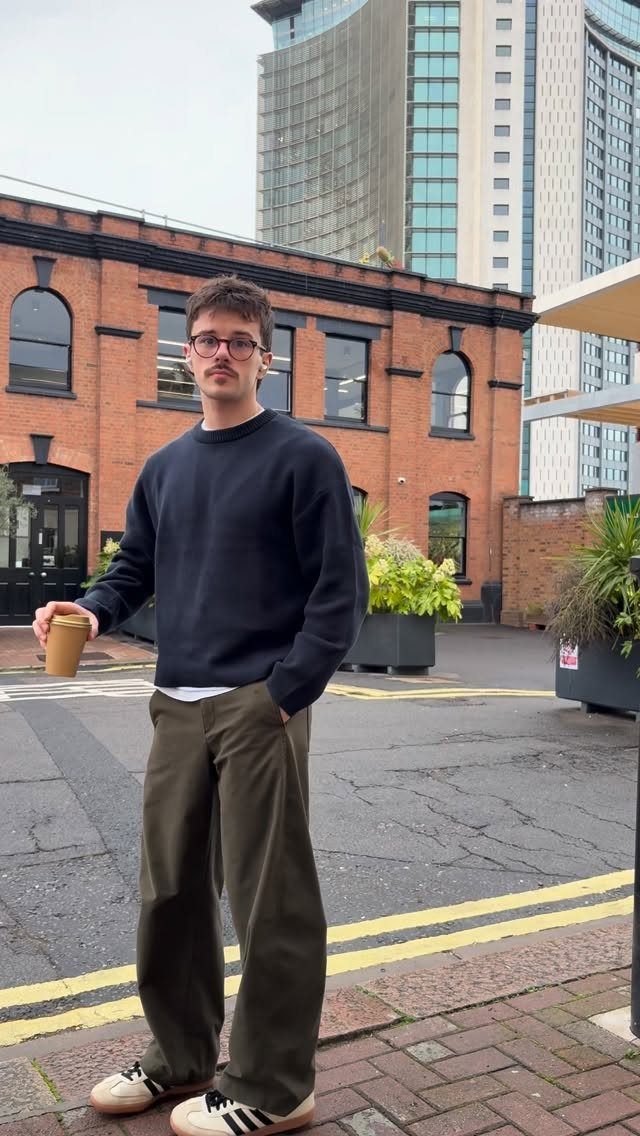 A man in a navy sweatshirt and olive trousers standing outdoors holding a coffee cup.