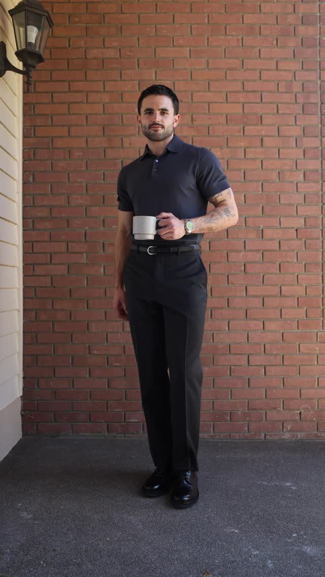 Man in a dark polo shirt and black tailored trousers standing against a brick wall.