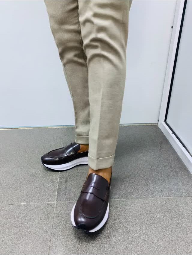 Dark brown leather loafers with white soles worn with tan trousers on a grey floor.