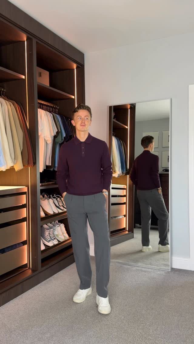 Man in a purple polo and grey trousers posing in a modern walk-in closet.