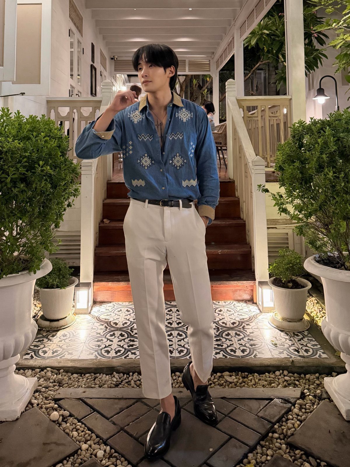 Man in a blue patterned shirt and white trousers standing on a tiled porch.