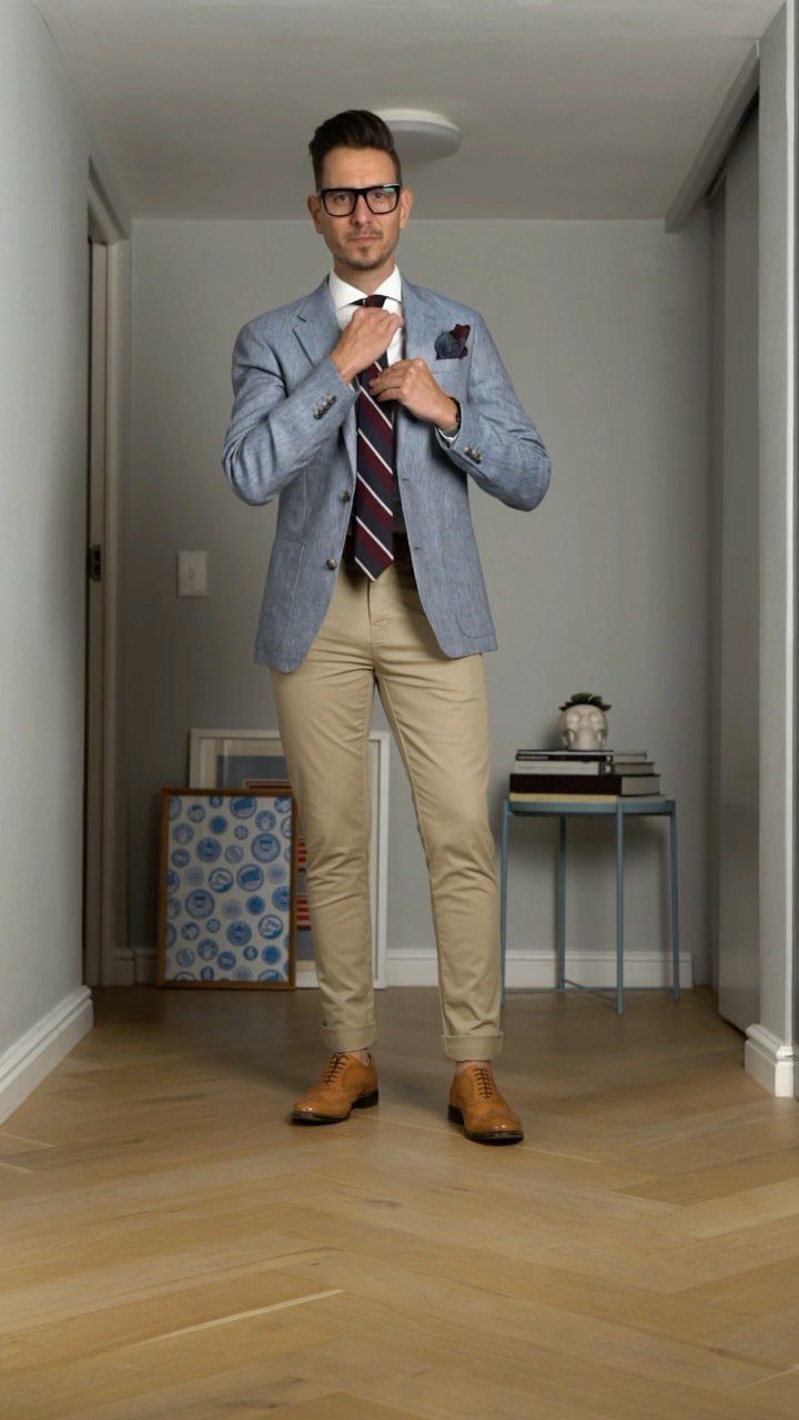 Man in a light blue blazer, striped tie, and tan slim-fit chinos standing in a hallway.