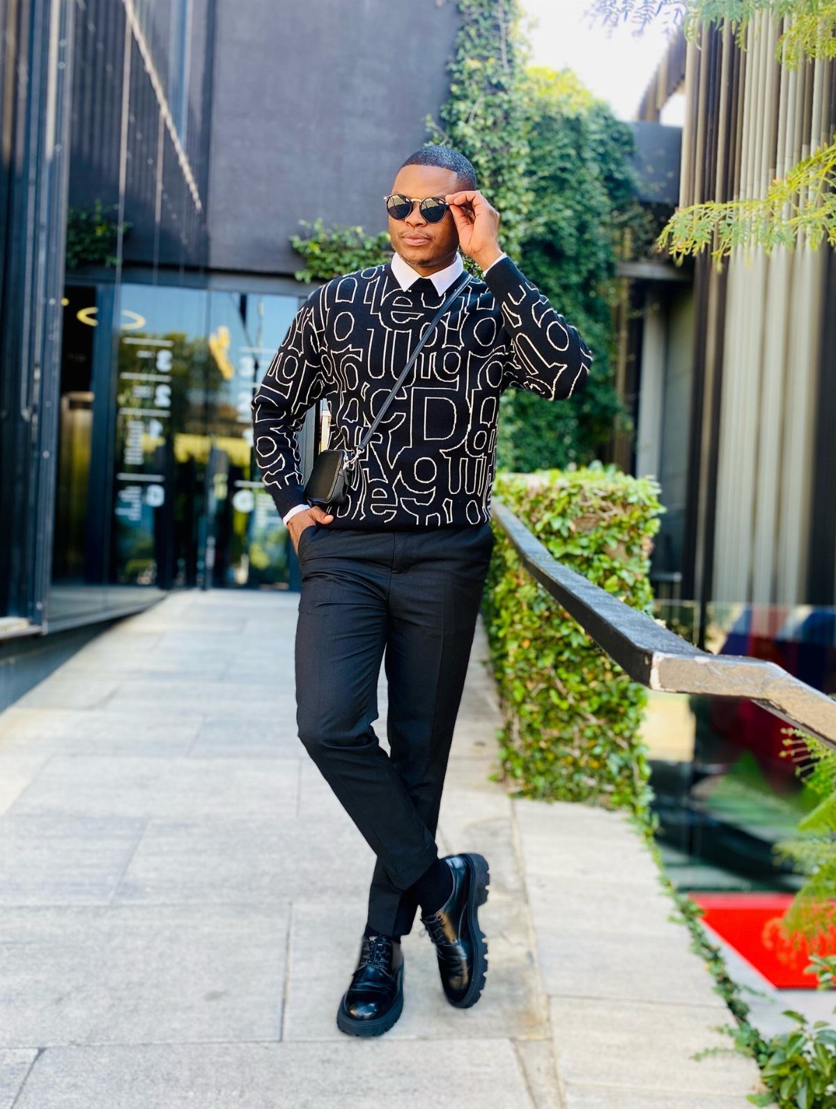 Man in a graphic sweater, collared shirt, black trousers, and loafers standing outdoors.