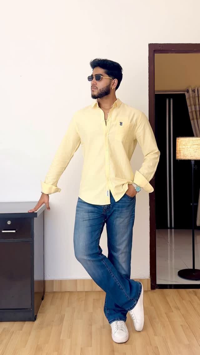 Man in a yellow shirt and blue jeans posing against a white wall.
