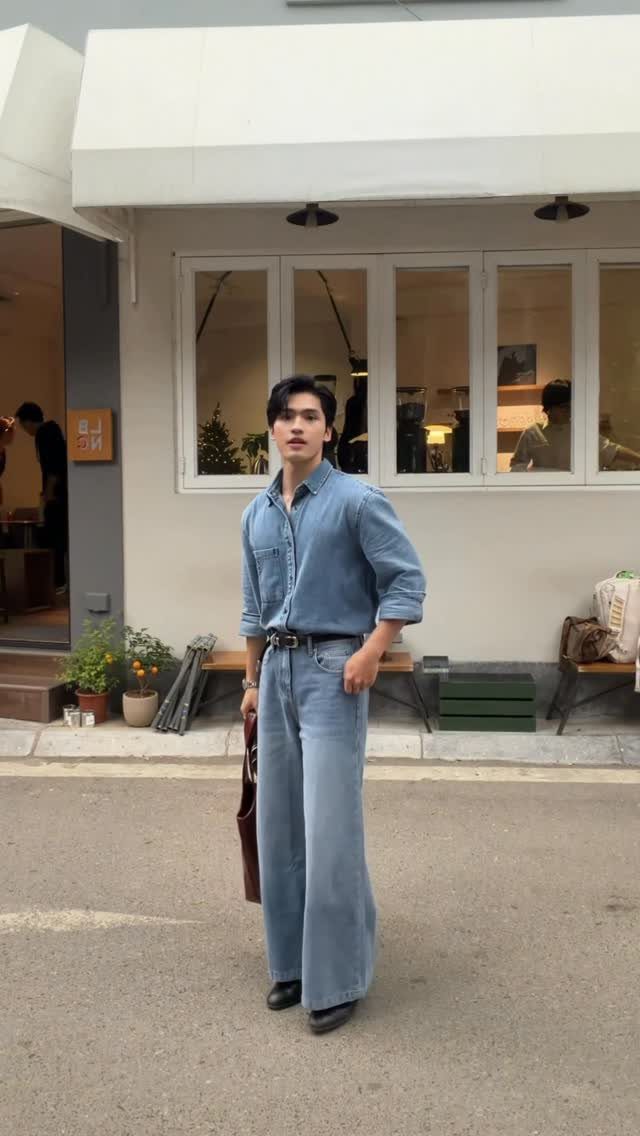 Man in denim shirt and wide-leg jeans standing in front of a white building.