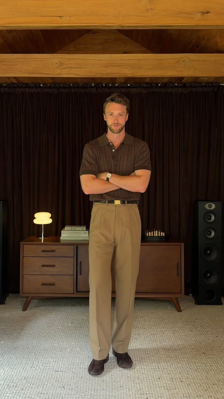Man standing in a brown polo shirt and tan trousers against dark curtains.