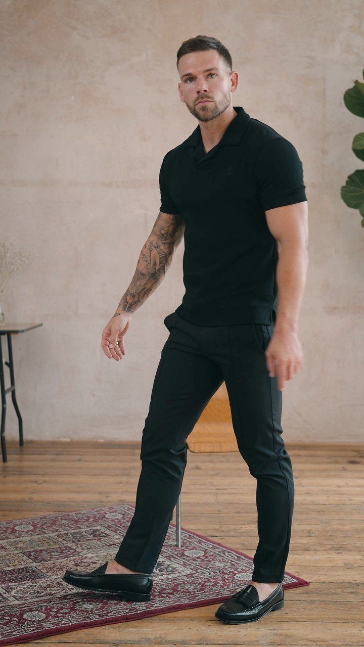 Man posing in a black polo shirt, black trousers, and black loafers on a rug.