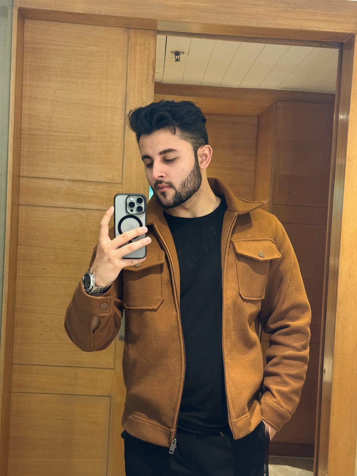 Man in a brown textured jacket and black t-shirt taking a mirror selfie.