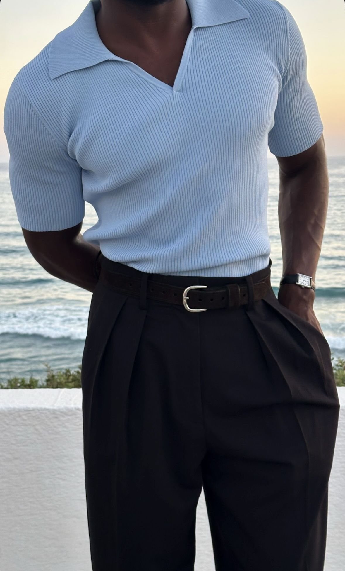 Man wearing a light blue polo shirt tucked into dark brown pleated trousers.