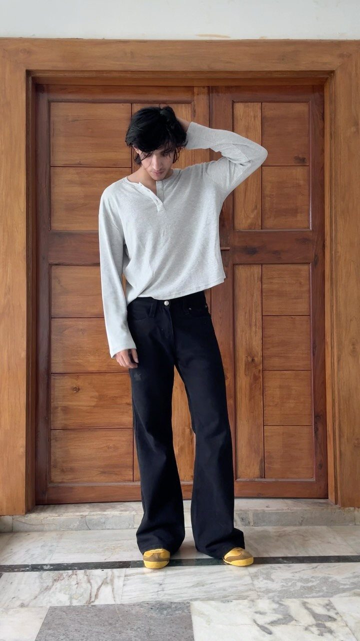 A man wearing a white long-sleeved henley shirt and black wide-leg trousers standing before a wooden door.