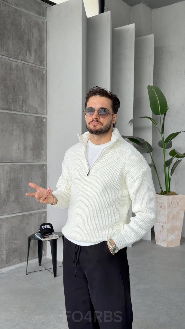 A man posing in a white textured quarter-zip knit sweater and black trousers.