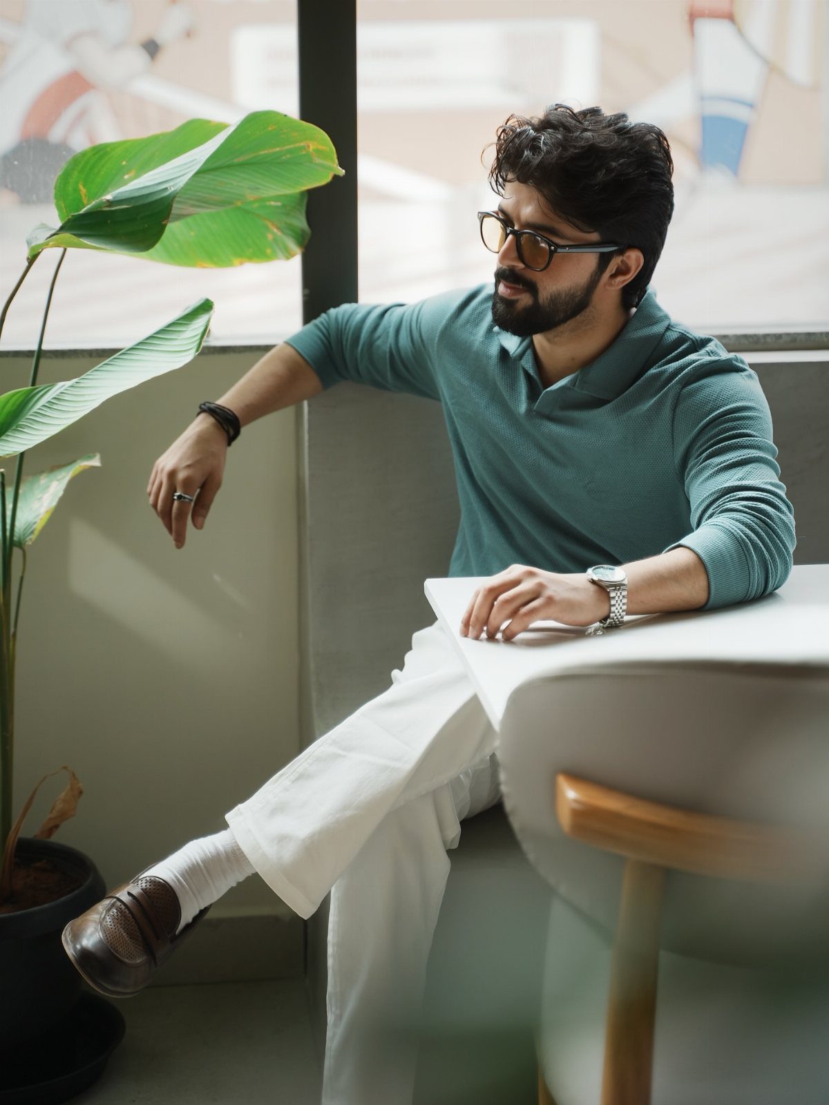 A man wearing a teal long-sleeved polo shirt and white trousers sitting by a plant.