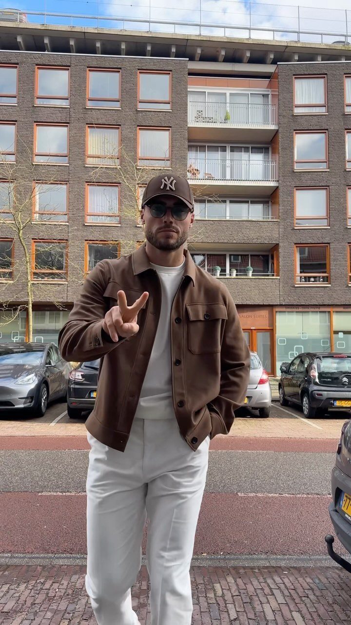 Man wearing a brown utility jacket over a white sweater and white trousers standing on a city street.