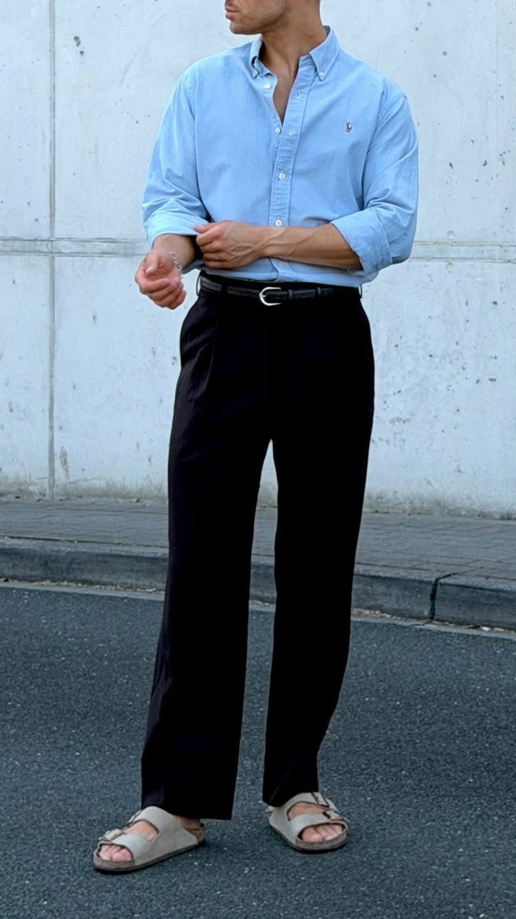 Man wearing a light blue button-down shirt, black trousers, and beige sandals standing outdoors.