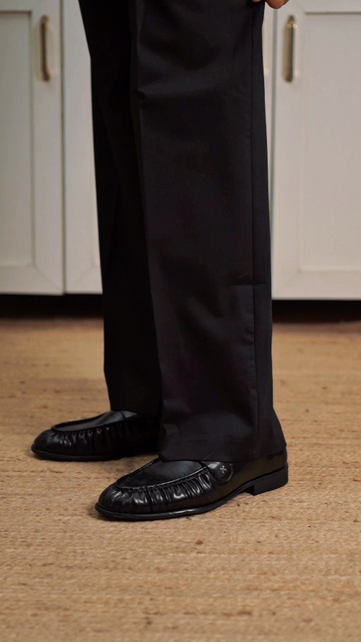 Black leather loafers worn with black trousers on a tan textured carpet.