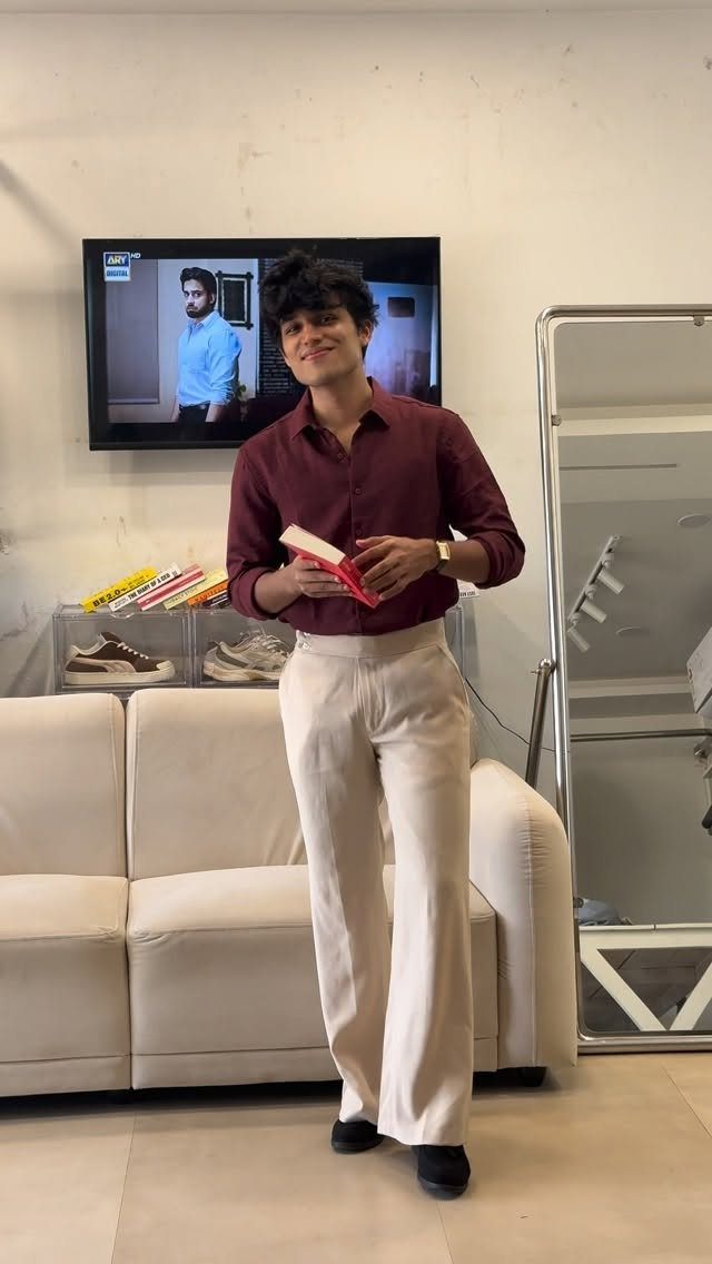 A smiling man wearing a maroon shirt and cream wide-leg trousers holding a red book.