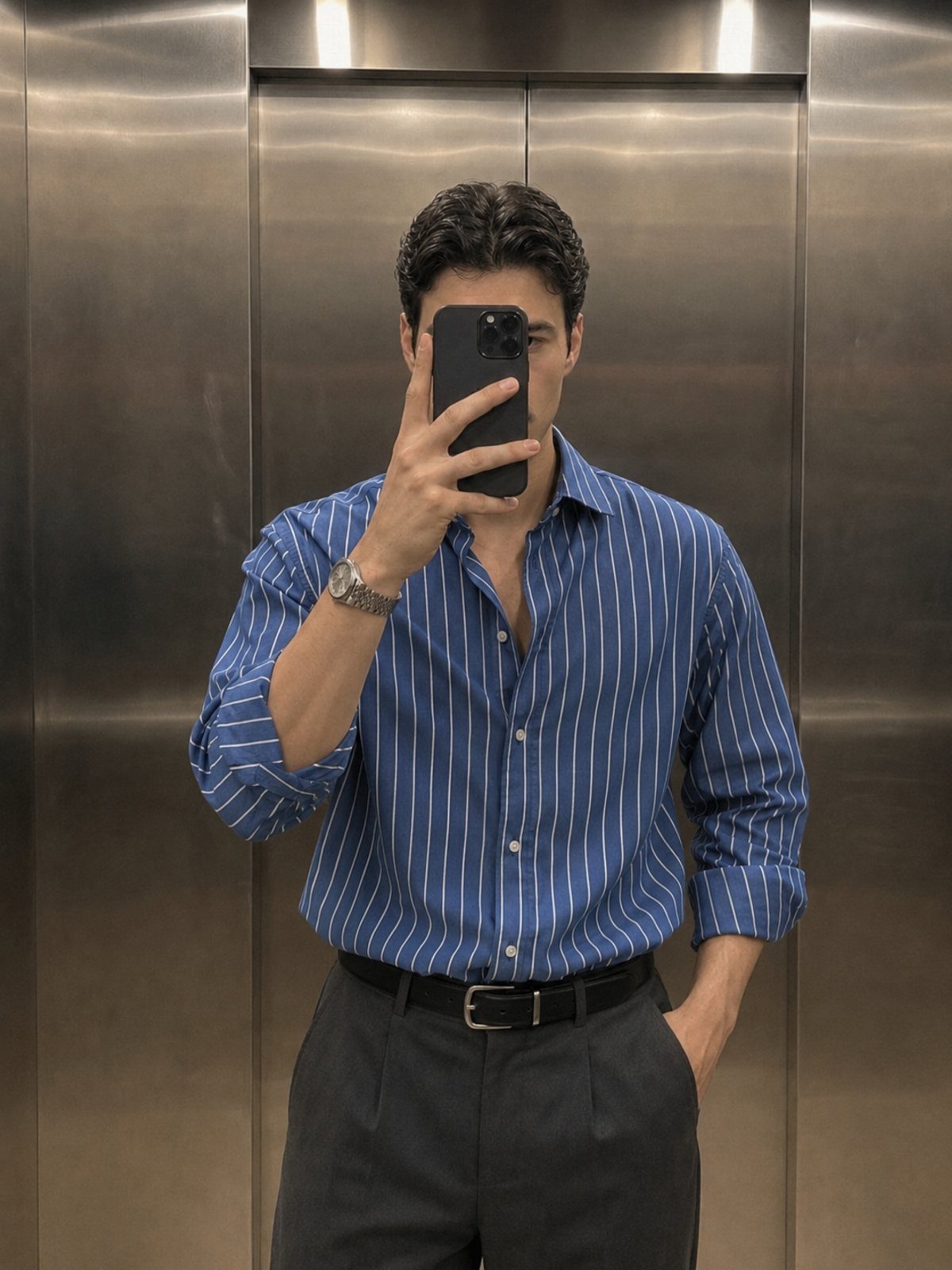 Man in a blue striped button-down shirt and grey pleated trousers taking a mirror selfie.