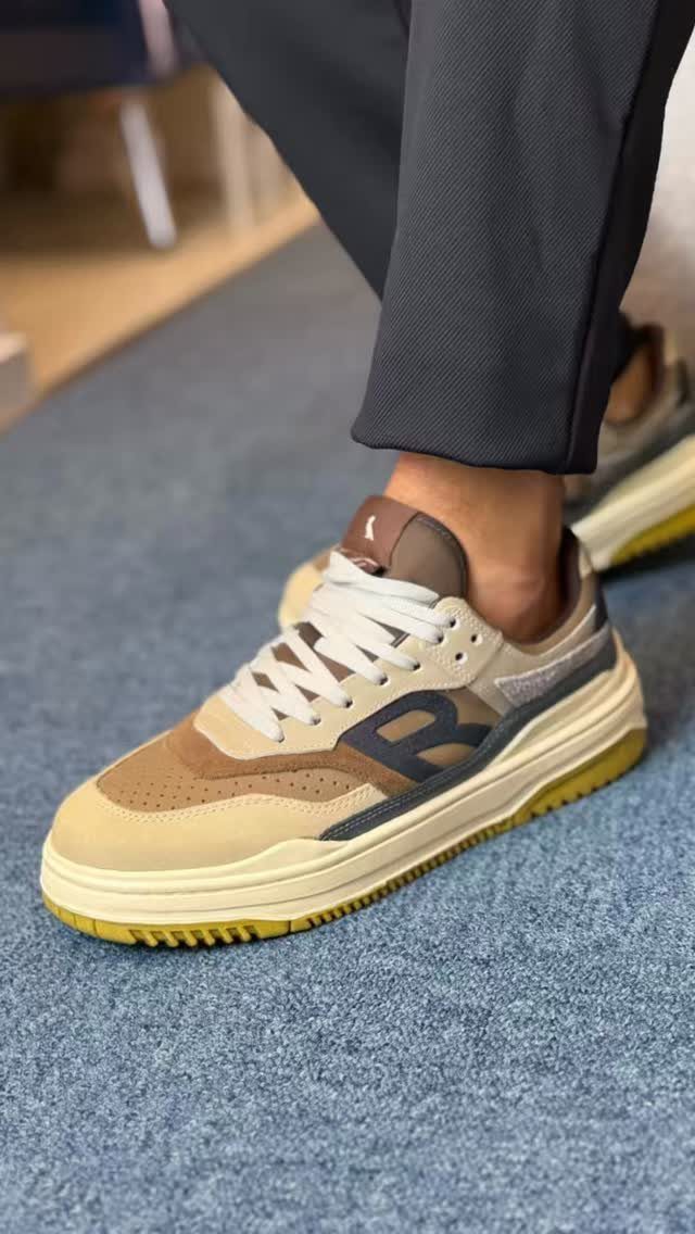 Tan and brown suede sneakers with white laces worn by a person in dark trousers.