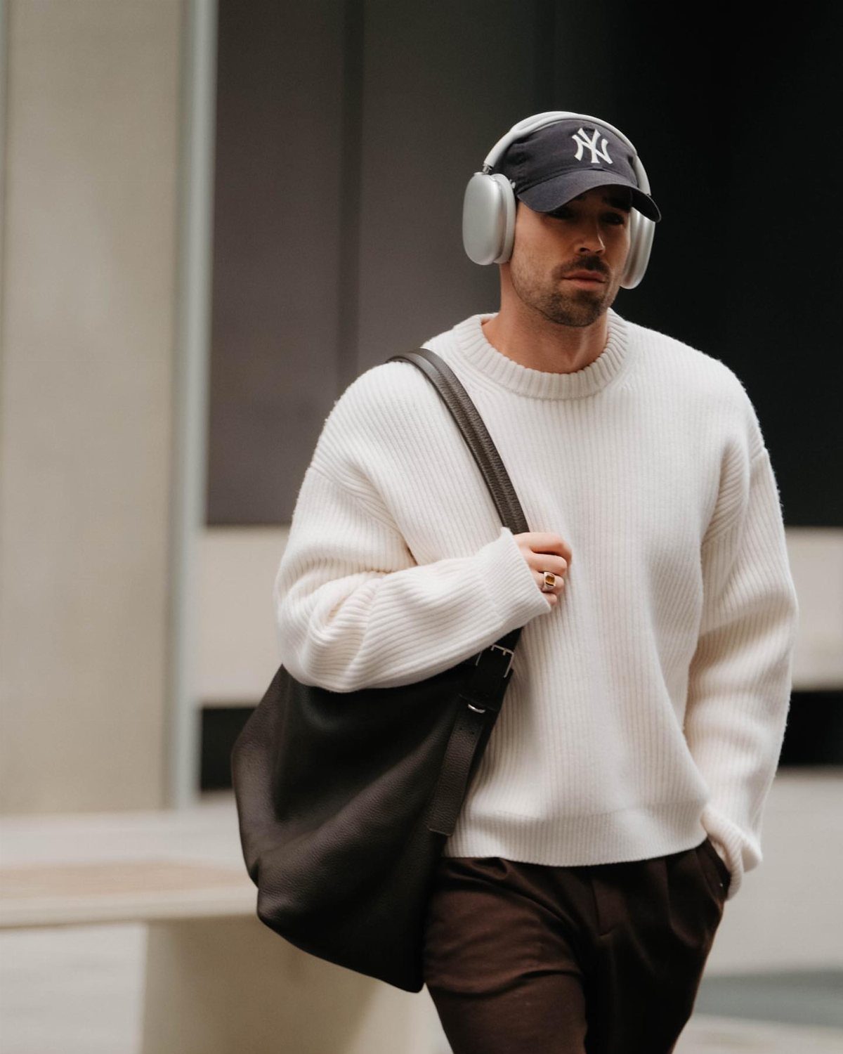 Man in a white ribbed sweater and dark trousers walking with a large black bag.