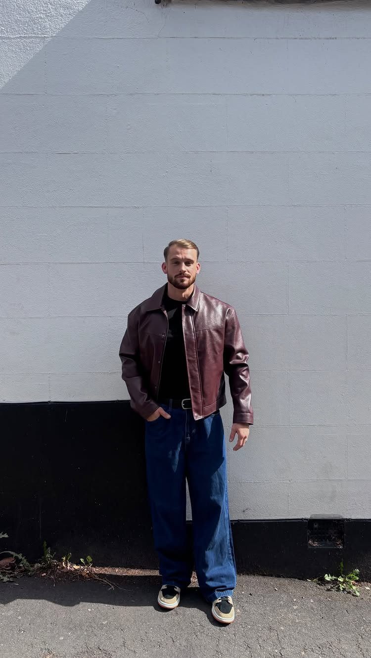 A man posing in a maroon leather jacket and wide blue jeans against a grey wall.