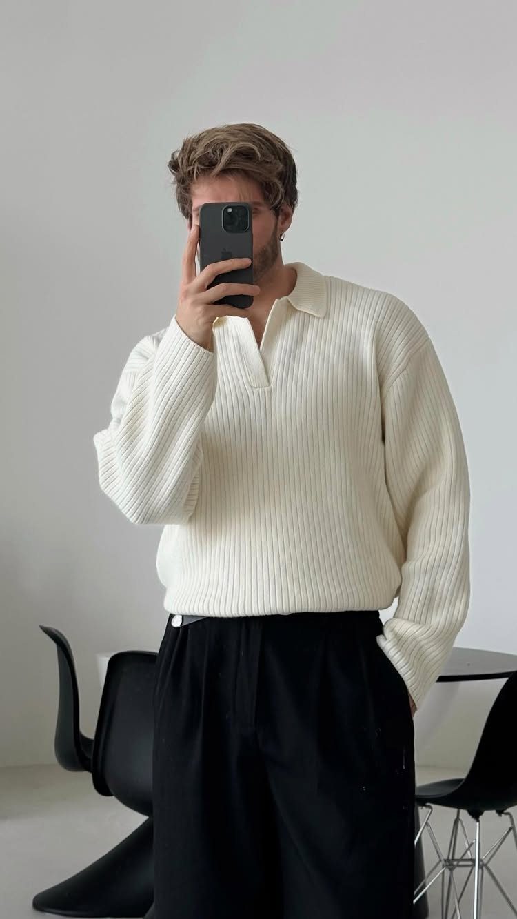 Man in a cream ribbed polo sweater and black pleated trousers taking a mirror selfie.