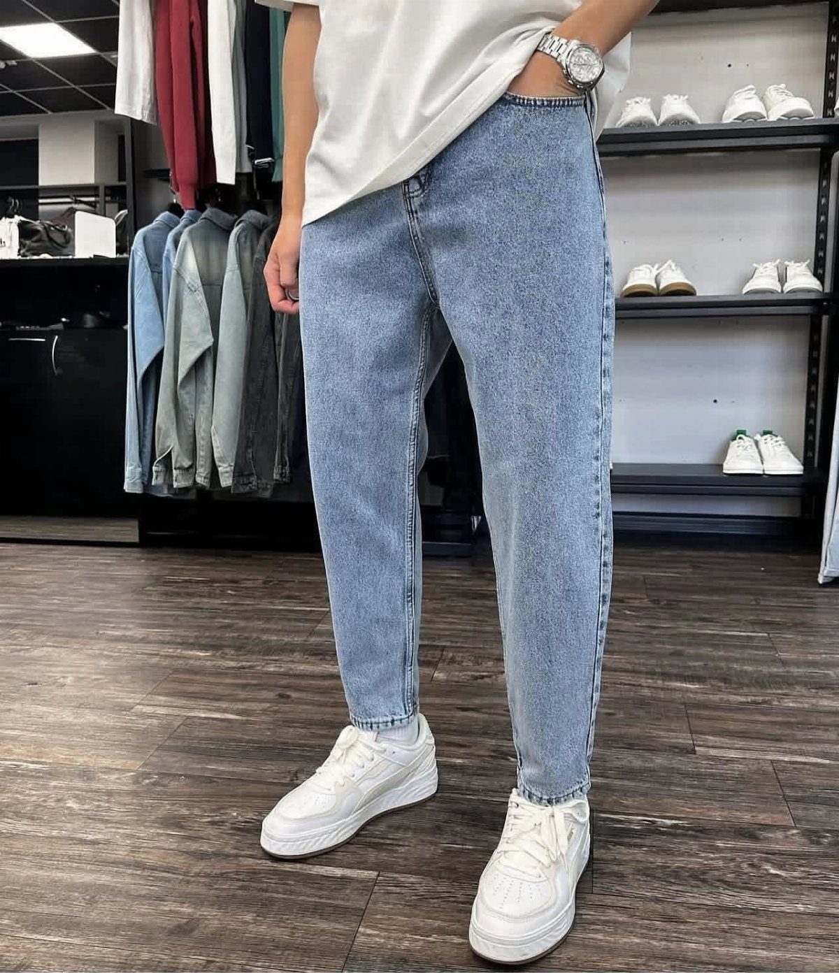 Man wearing light blue tapered denim jeans, a white t-shirt, and white sneakers in a clothing store.