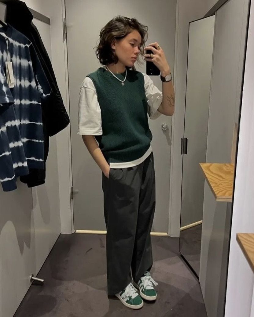 Woman in a white t-shirt, dark green sweater vest, and gray trousers posing in a mirror.