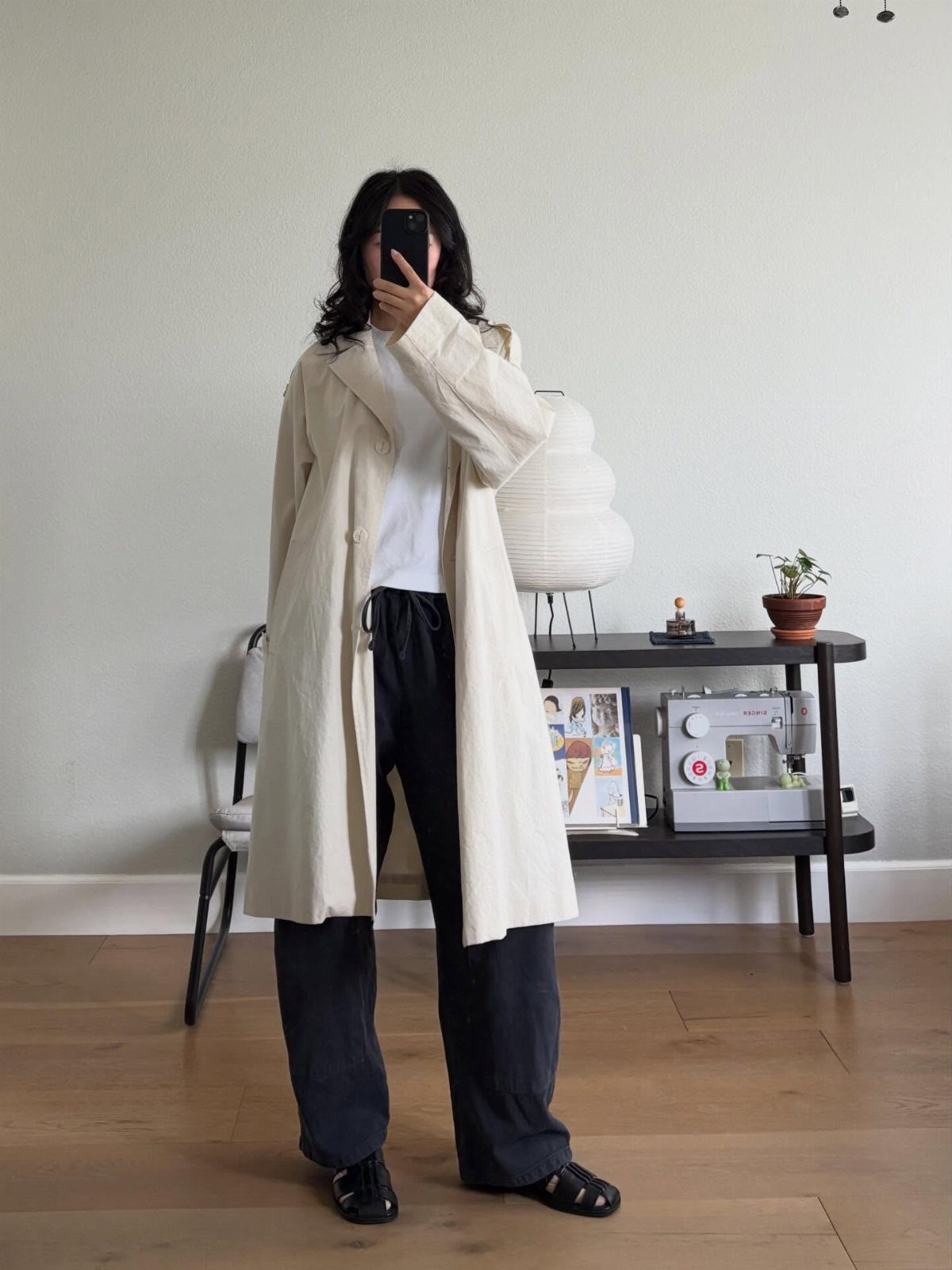 Person wearing a long cream coat, white shirt, and black wide-leg trousers standing indoors.