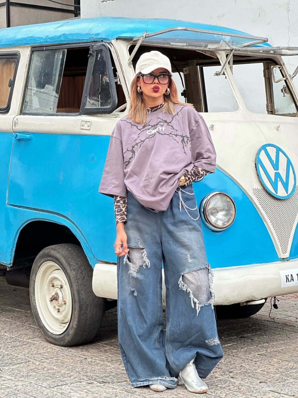 Woman wearing an oversized lavender graphic tee and baggy ripped blue jeans standing by a vintage van.