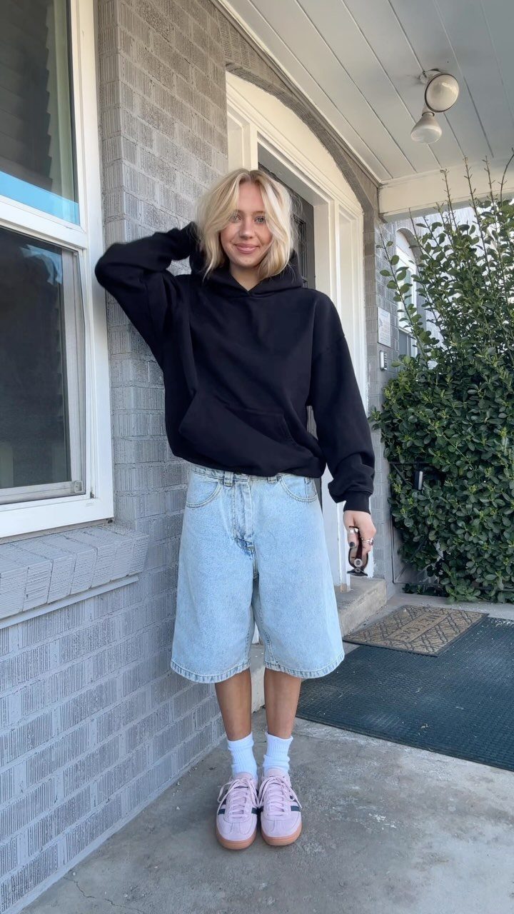 Blonde woman wearing a black oversized hoodie and light blue denim shorts standing outdoors.