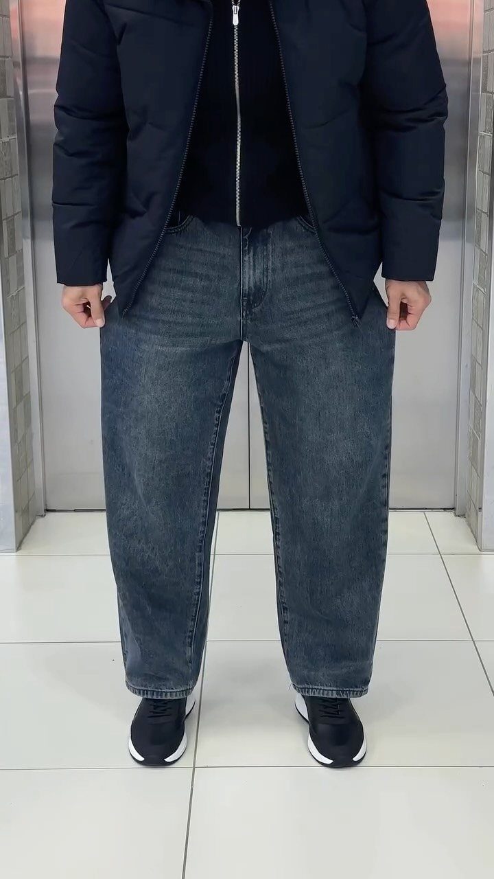 Person wearing a dark puffer jacket, black zip-up shirt, blue jeans, and black sneakers.