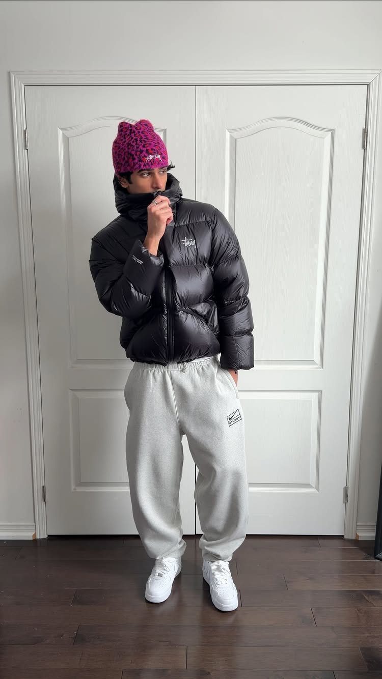 A man wearing a black puffer jacket, grey sweatpants, and a pink beanie.