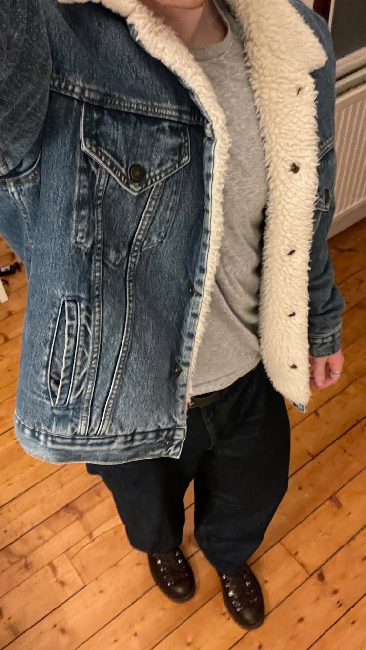 A person wearing a denim sherpa jacket, grey shirt, dark pants, and brown boots.