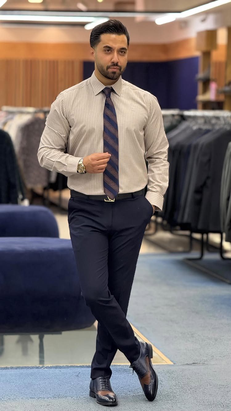 Man posing in a striped shirt, dark tie, navy trousers, and black loafers.