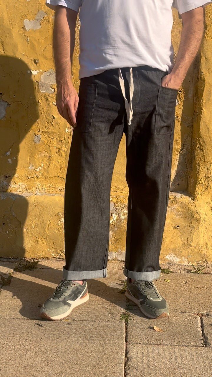 Man standing against a yellow wall wearing a white t-shirt, dark grey drawstring trousers, and grey sneakers.