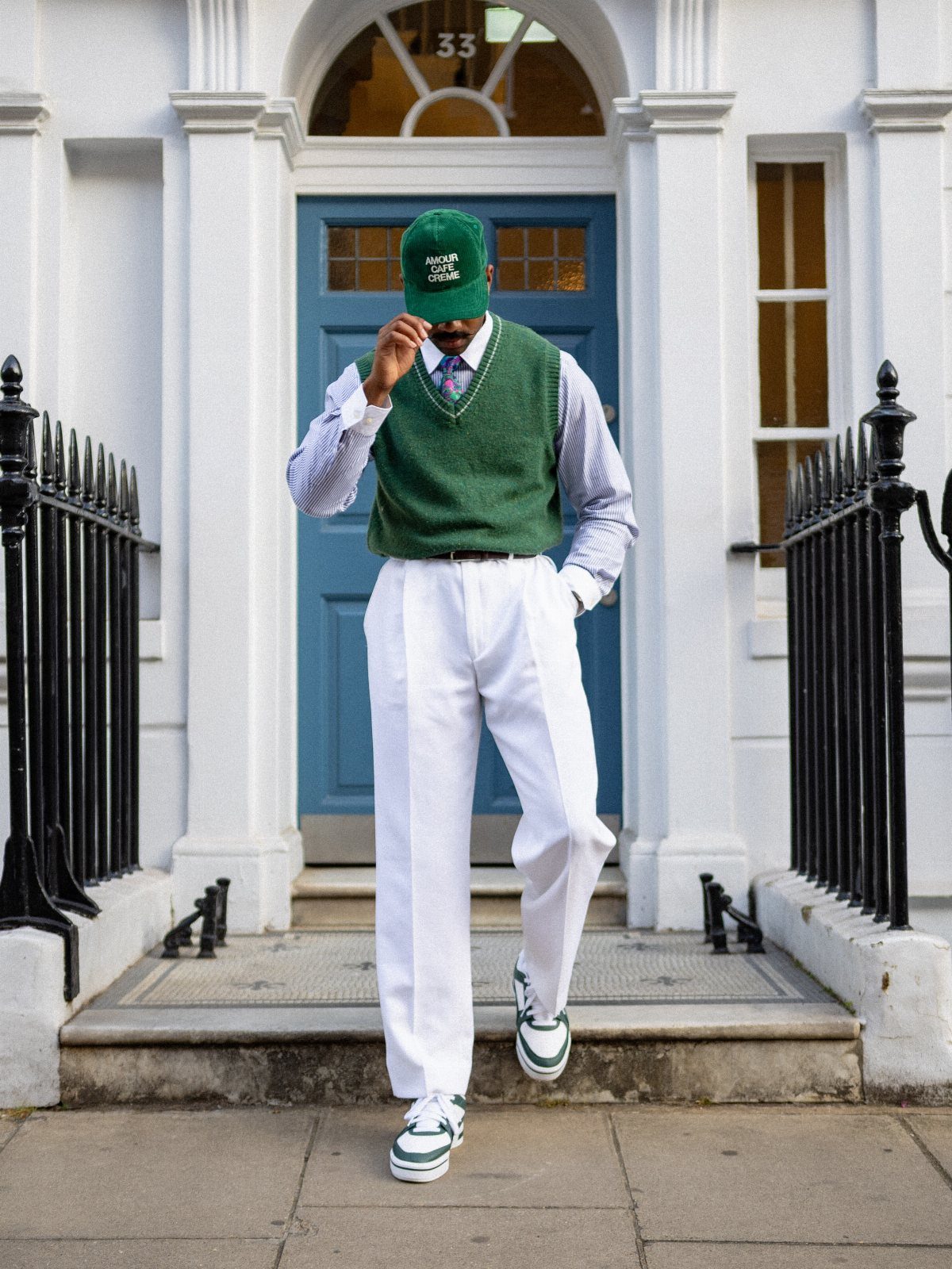 Man in green sweater vest, white trousers, and green cap walking down steps in front of a blue door.
