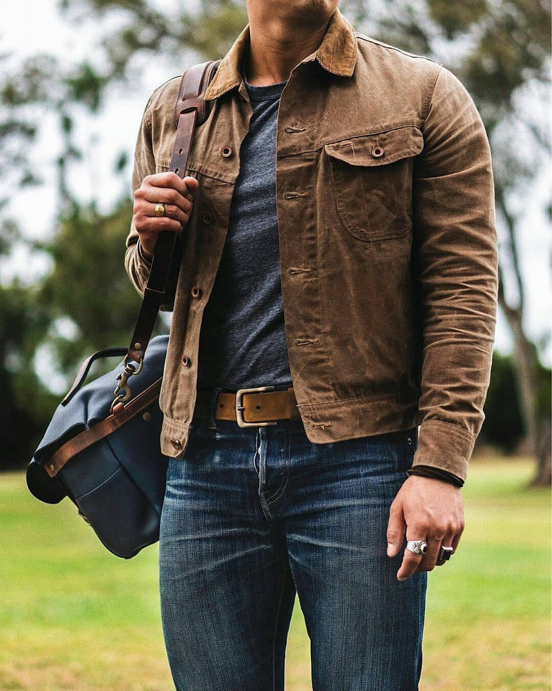 Man wearing a brown suede jacket, grey t-shirt, and blue denim jeans with a leather bag.