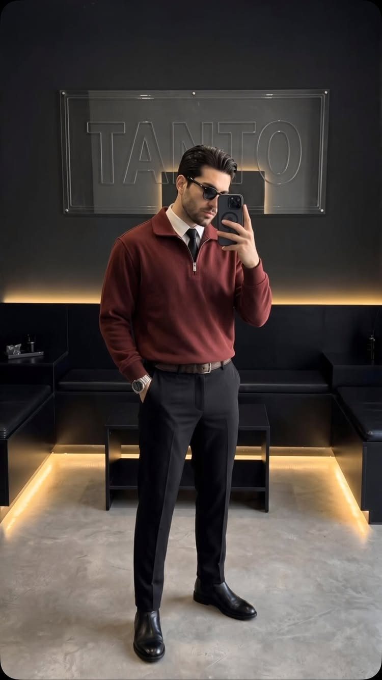 Man in a maroon pullover, white shirt, tie, and black trousers posing for a mirror selfie.
