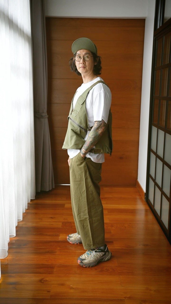 Man posing in olive green utility vest, matching trousers, and a green cap indoors.