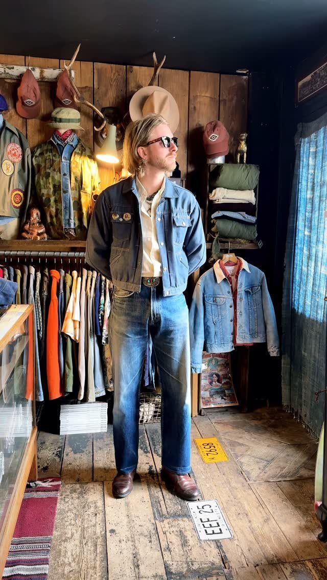 Man in denim jacket and jeans standing inside a rustic vintage clothing store.