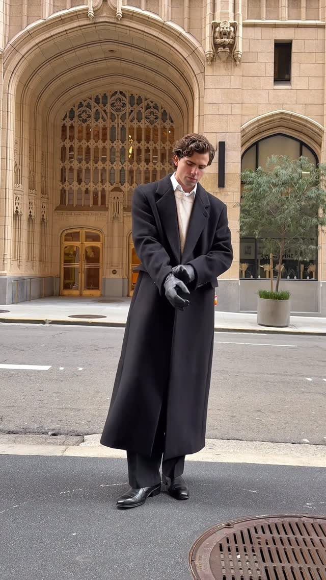 A man wearing a long black overcoat and gloves standing on a city street.
