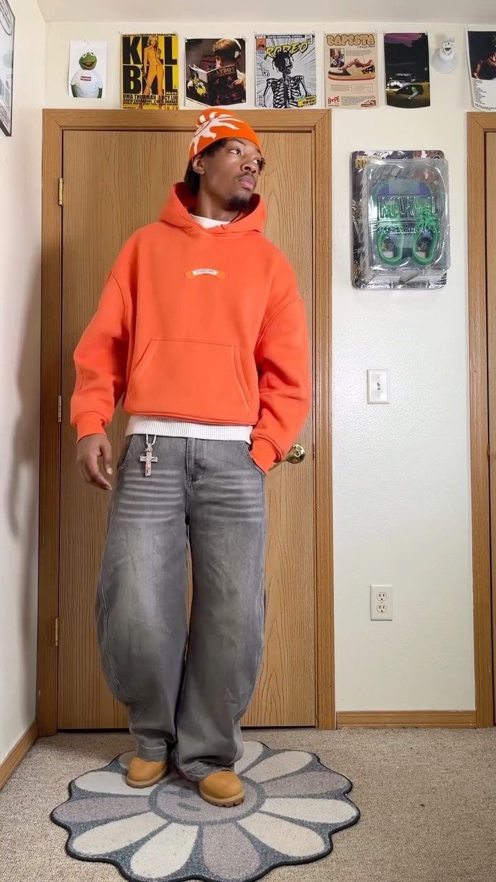 A man wearing a bright orange hoodie, orange beanie, and baggy grey jeans standing indoors.