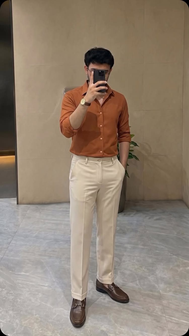 A man posing in a burnt orange shirt, cream trousers, and brown leather shoes.